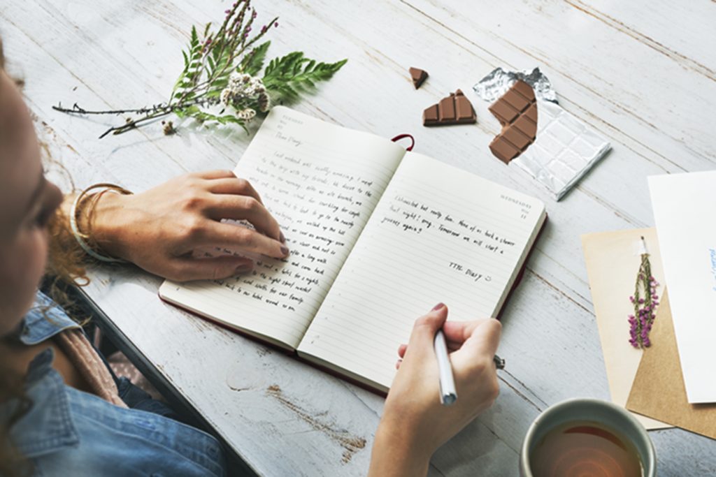 A person writes in a notebook on a wooden table. There are pieces of nature on the table.