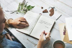 A person writes in a notebook on a wooden table. There are pieces of nature on the table.