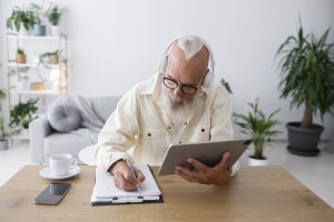 Neat-looking man with white hair and interesting-looking beard sits in a light-filled room with plants behind him. He has headphones on and is writing.