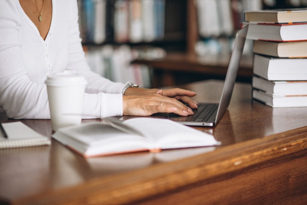 Woman works at laptop on a lovely wooden desk with books on the desk an on shelves behind her.
