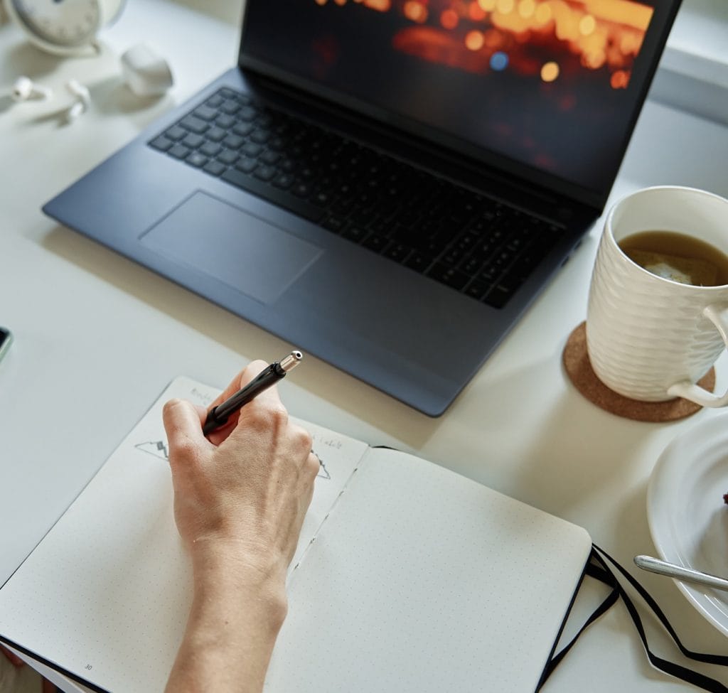 Taken from above - an open laptop sits on a desk, a mug of coffee beside it, a hand writing in a notebook in front of the laptop.