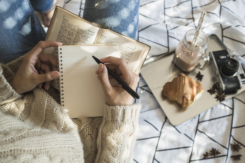 Woman with aran-type woollen sweater and jeans sits and writes in notebook with a hot chocolate, pastry and camera beside her. Cosy.