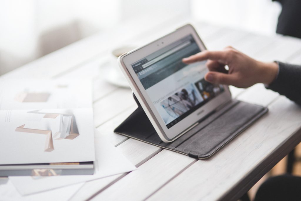 A person's hand using the touch screen on a tablet which is on a painted white table. A large photographic book lies beside the tablet on the table.