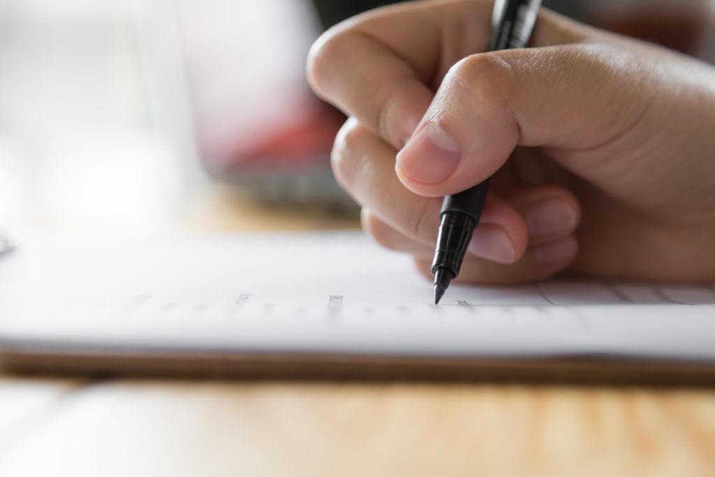 A woman's hand writes with a pen on some paper on a wooden desk.